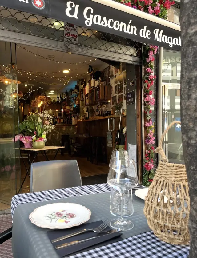 Entrada de El Gasconín de Magalí en la calle Gascona: una mesa con mantel de cuadros y plato de flores en primer plano, y la barra iluminada al fondo