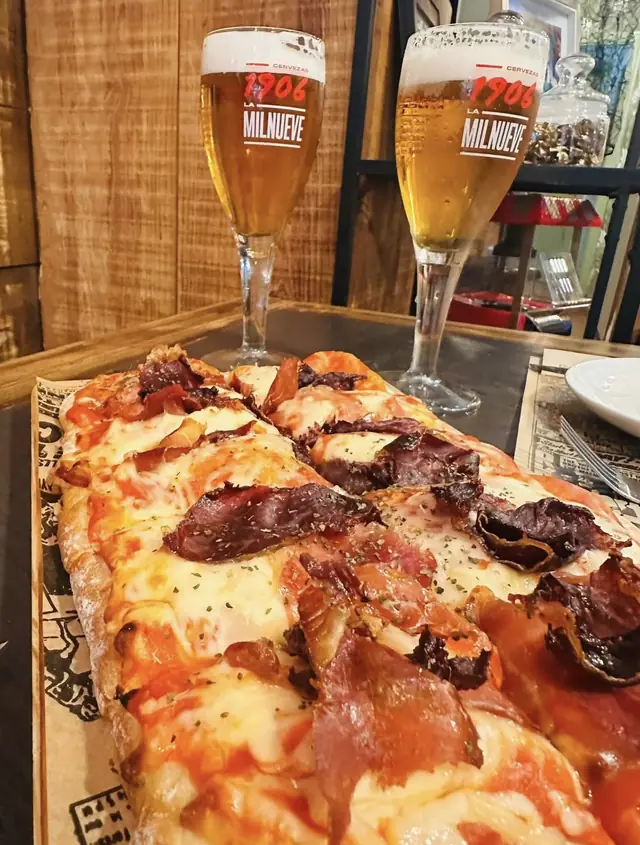 Pizza rectangular con jamón y cecina en tabla de madera, con dos cervezas al fondo